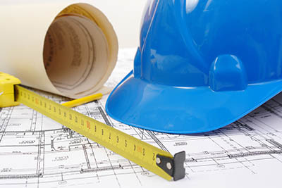 Closeup of blue hard hat, yellow pencil, measuring tape and building plans.